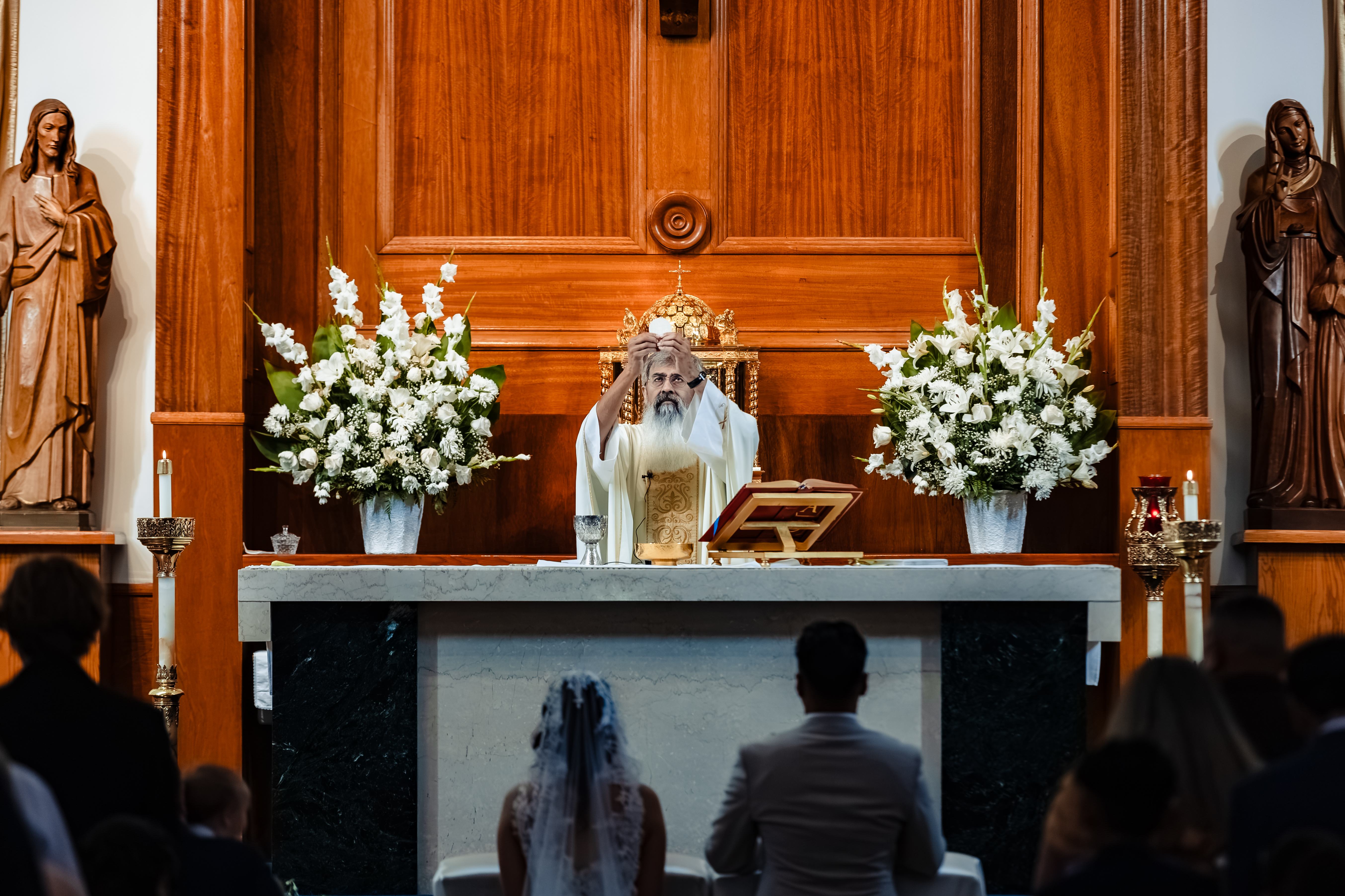 A priest elevates the Host at the consecration during a Catholic nuptial Mass at St. Anne's in Santa Ana, framed by the kneeling bride and groom