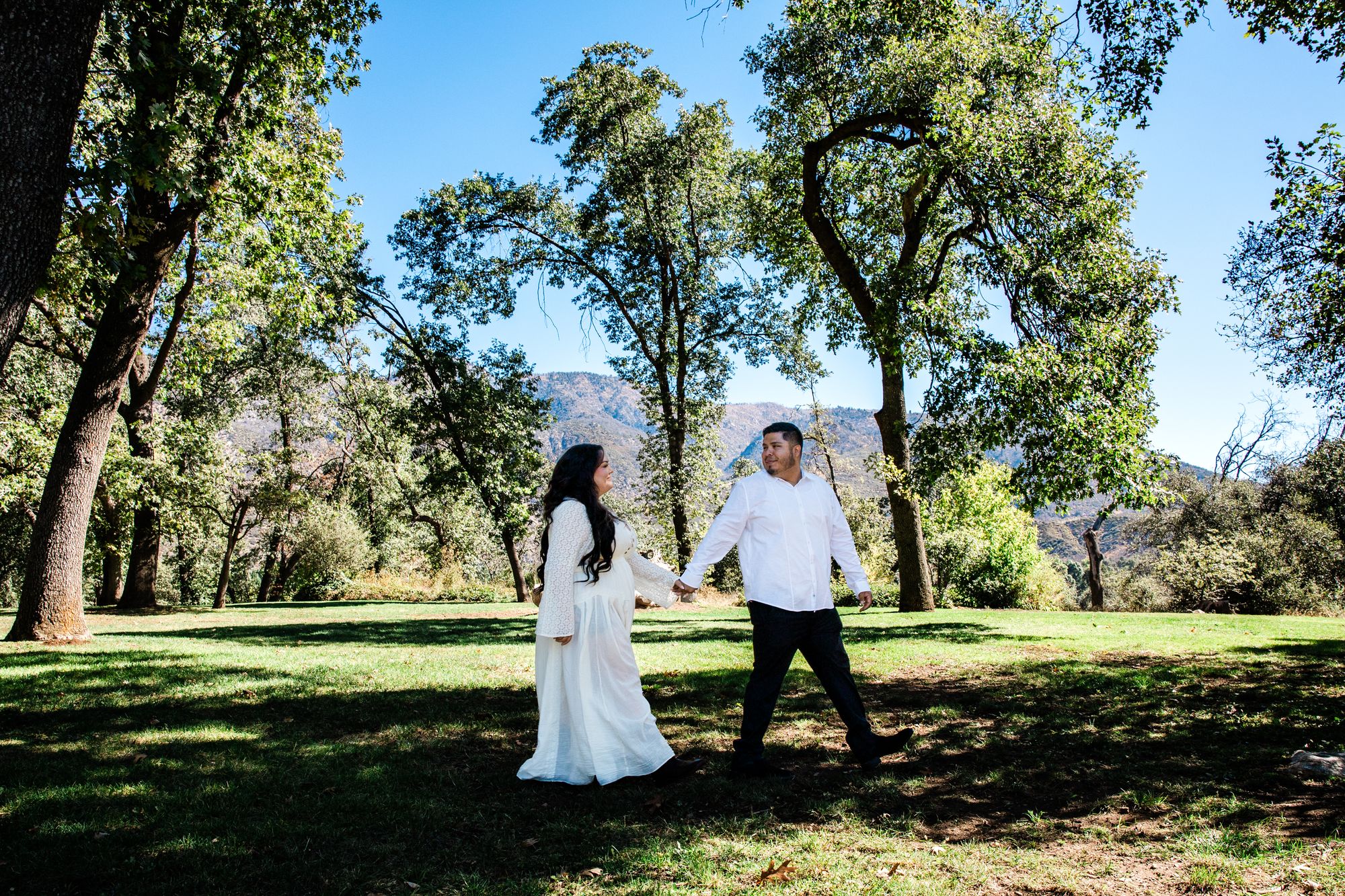 Eric and Cristina walking across a sunlit meadow ringed by oaks and mountains in Oak Glen, California