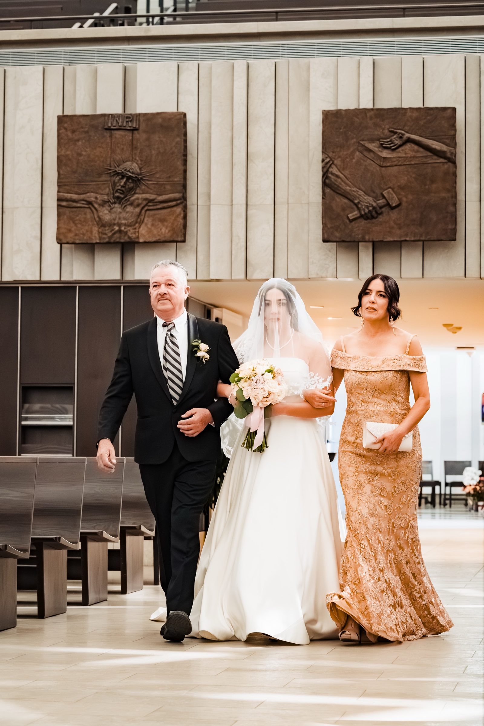 A bride walked down the aisle by both parents beneath the bronze Stations of the Cross at Christ Cathedral