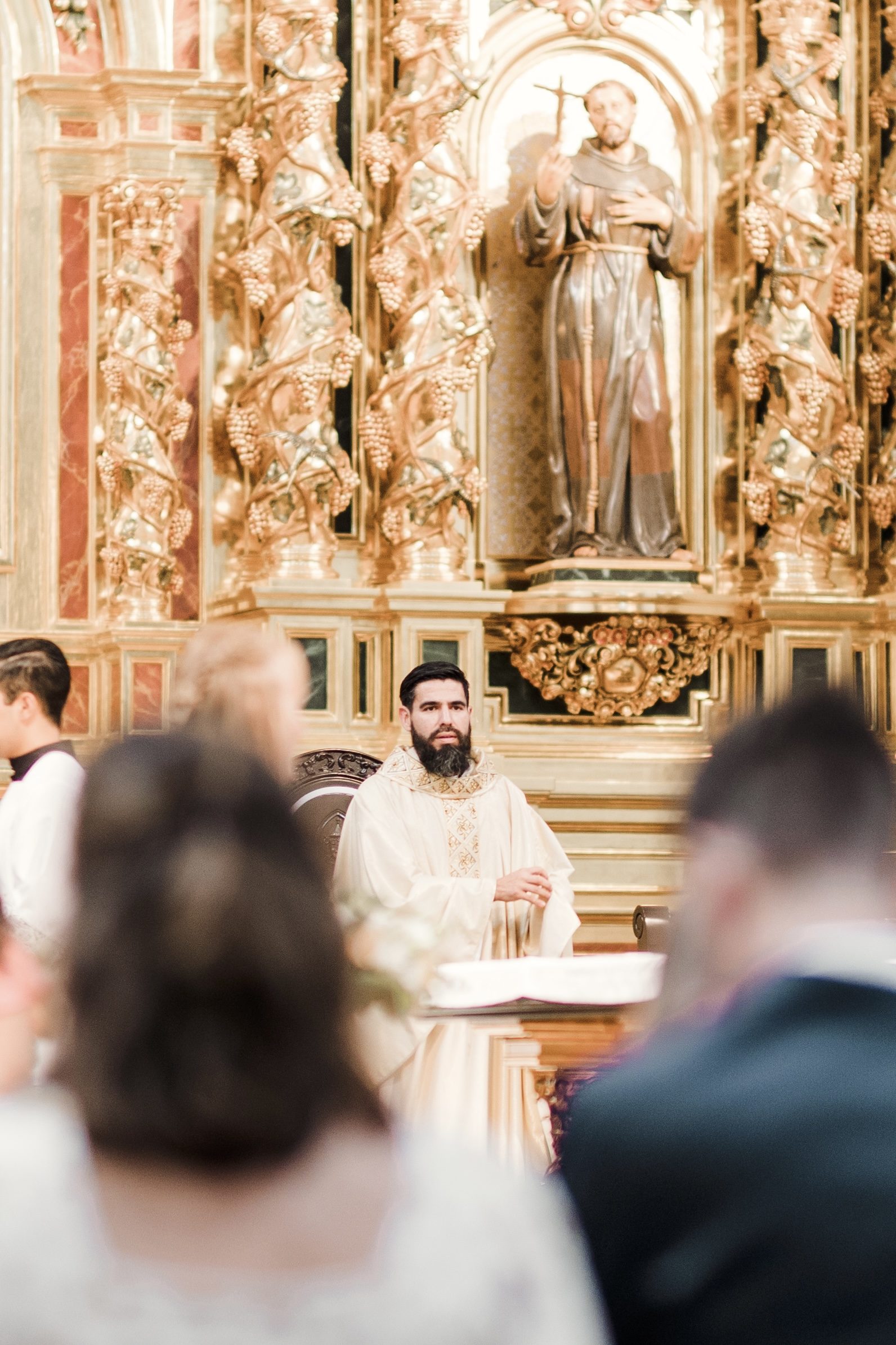 The celebrant seated beneath St. Francis in the gilded retablo at Mission Basilica San Juan Capistrano