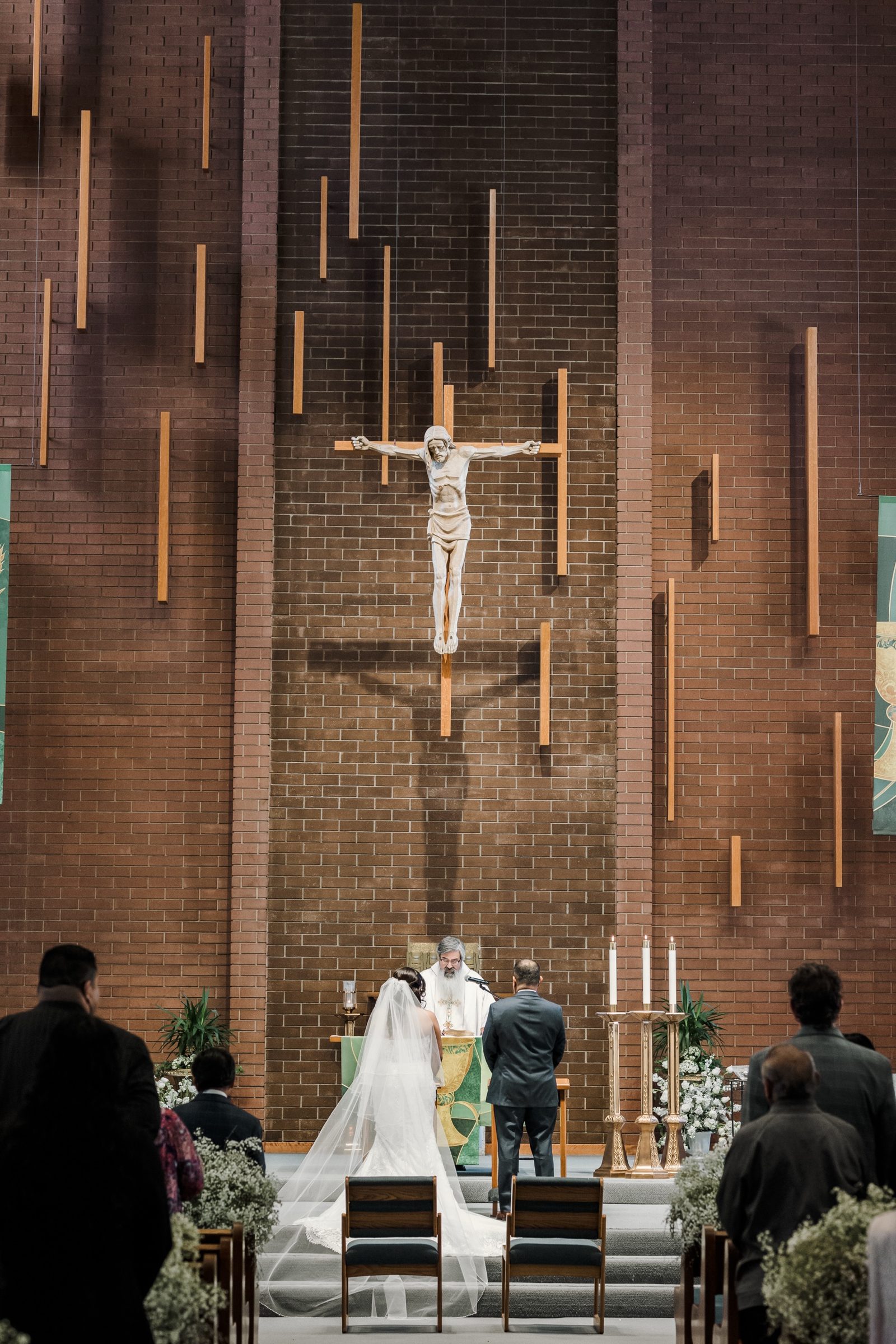 A bride and groom before the priest beneath the brick wall and crucifix of St. Angela Merici, Brea