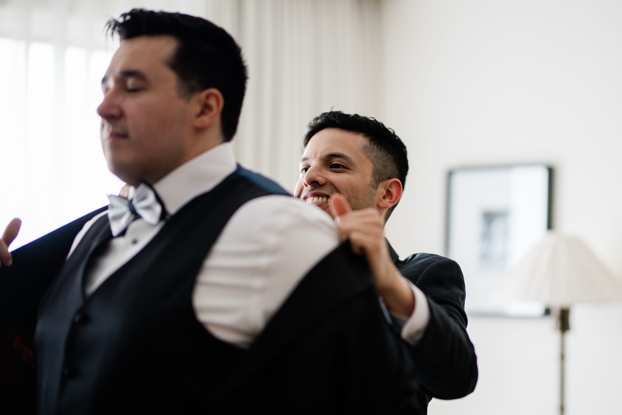 A groomsman helping Andrew into his tuxedo jacket