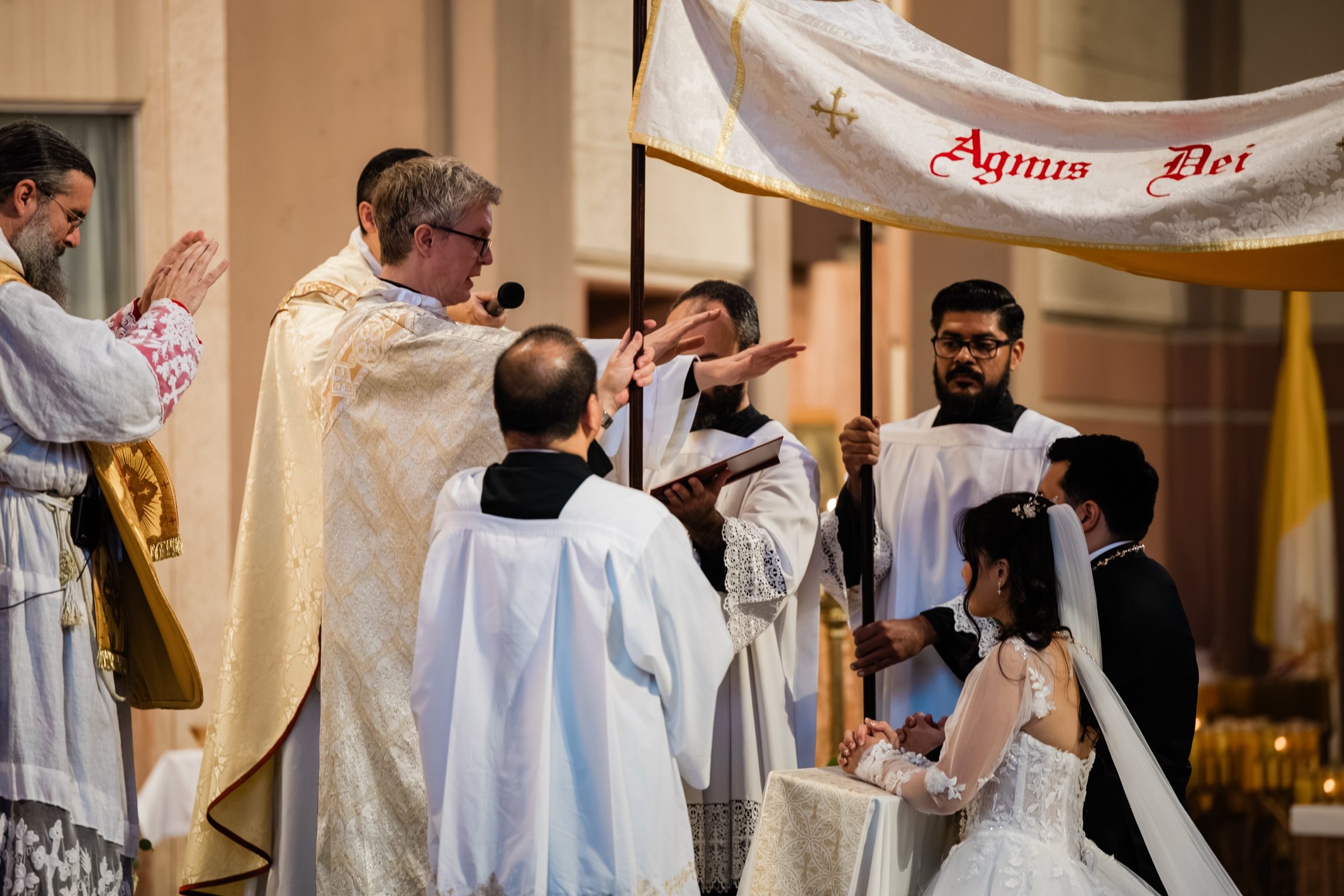 The nuptial blessing beneath a canopy embroidered Agnus Dei as priests extend their hands over the couple