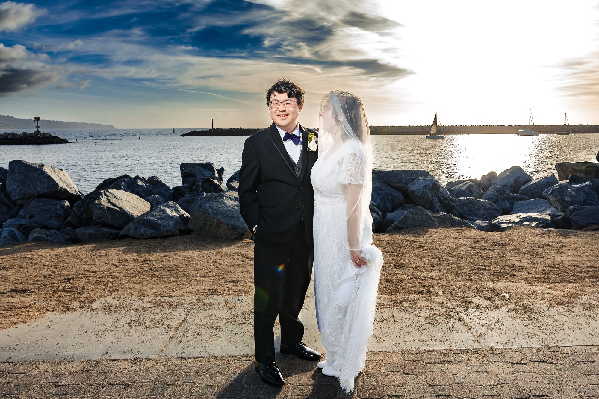 Bride and groom golden hour portrait at the King Harbor breakwater in Redondo Beach with a sailboat on the water